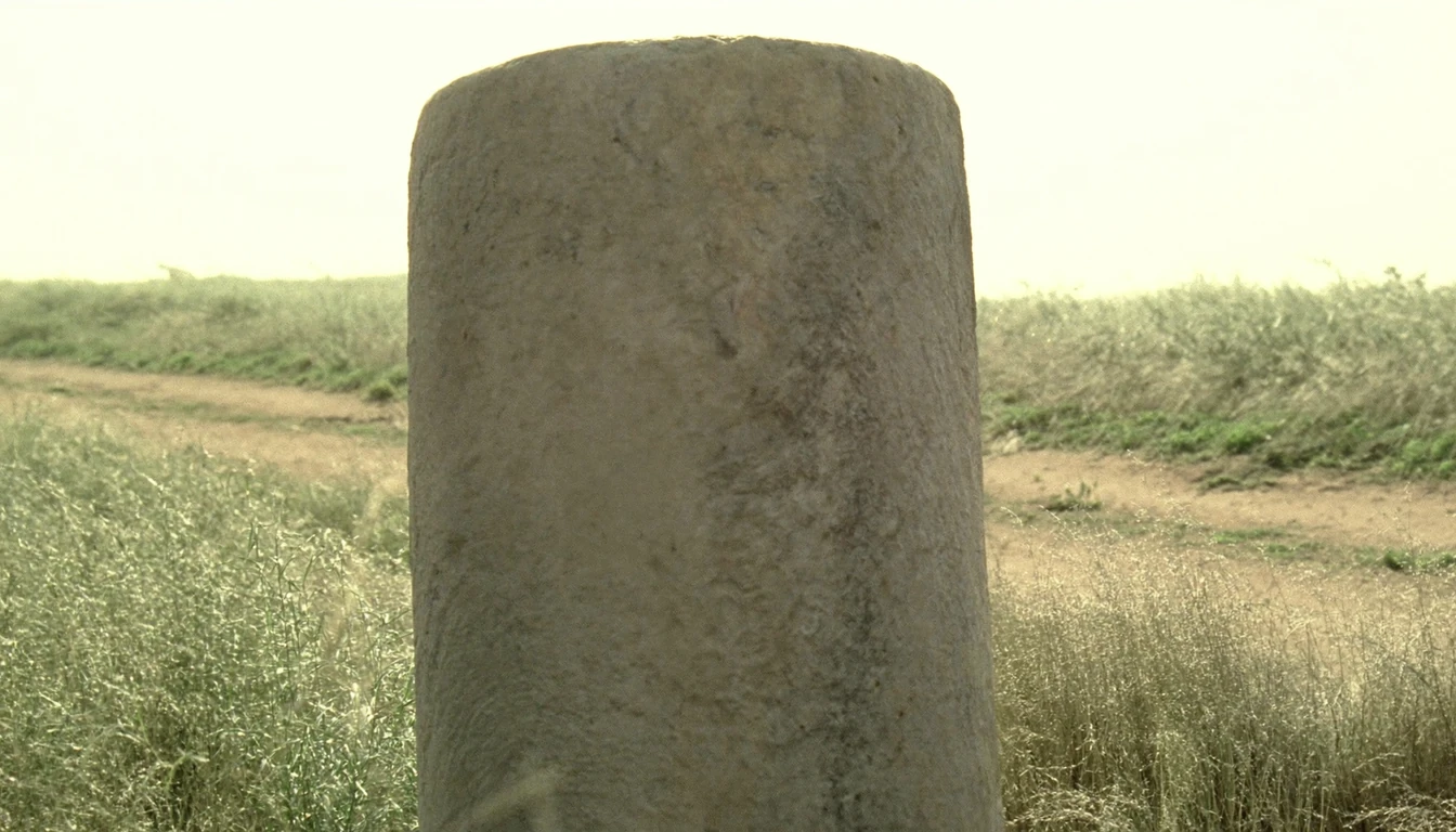 A plain uninscribed cylindrical stone milestone standing in dry grass, a dirt track running past it on both sides